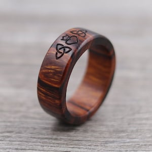 May include: A wooden ring with a Celtic knot and heart designs carved into the band. The ring is made of dark brown wood with a reddish hue.