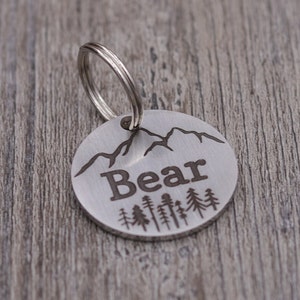 Könnte beinhalten: Silbernes Hundehalsband mit einer Bergkette und einer Reihe von Kiefern, die darauf graviert sind. Das Schild trägt die Aufschrift "Bear".