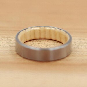 May include: A close-up of a ring with a brushed silver exterior and a light-colored wood interior. The ring is set against a light brown wooden surface, showcasing the contrast between the metal and wood.