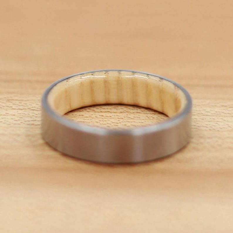 May include: A close-up of a ring with a brushed silver exterior and a light-colored wood interior. The ring is set against a light brown wooden surface, showcasing the contrast between the metal and wood.