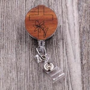 May include: A wooden badge reel featuring a cross and lily design. The round reel is made of wood with a laser-etched cross and lily. It has a clear plastic strap and a metal clip for attaching to clothing.