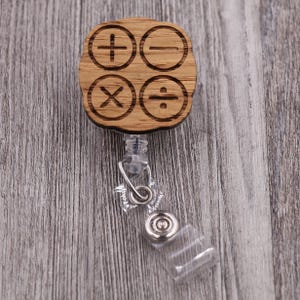 Peut inclure: Un enrouleur de badge en bois avec une finition marron clair. Le reel présente quatre symboles mathématiques : addition, soustraction, multiplication et division. Le reel a un cordon rétractable en plastique transparent et un clip en métal.