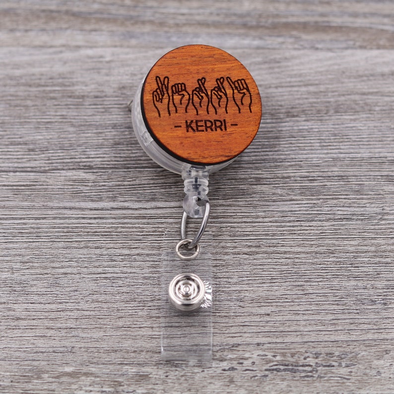 May include: A wooden badge reel with a brown wooden circle featuring the sign language spelling of the name "KERRI".