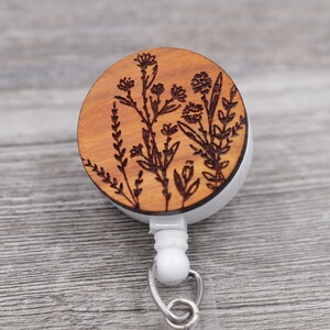 Puede incluir: Un carrete de identificaci&oacute;n de madera con un dise&ntilde;o floral. El carrete es marr&oacute;n con un cord&oacute;n retr&aacute;ctil blanco y un clip plateado.