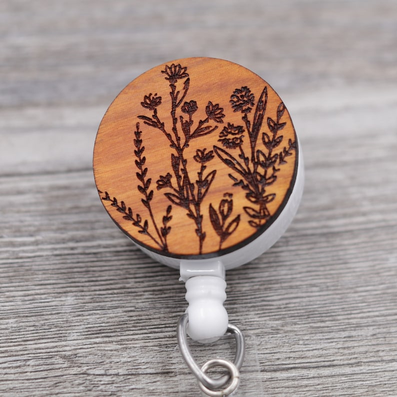 Puede incluir: Un carrete de identificaci&oacute;n de madera con un dise&ntilde;o floral. El carrete es marr&oacute;n con un cord&oacute;n retr&aacute;ctil blanco y un clip plateado.