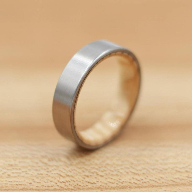 May include: A silver-colored ring with a flat, brushed metal exterior and a light-colored interior. The ring is set against a light brown wooden surface, showcasing its simple, modern design. This ring is a piece of jewelry.