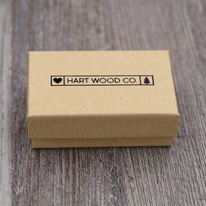 May include: A small, closed, rectangular cardboard box in a natural tan color. The box has a black stamp that reads "HART WOOD CO." with a heart and a tree symbol. The box is on a wooden surface.
