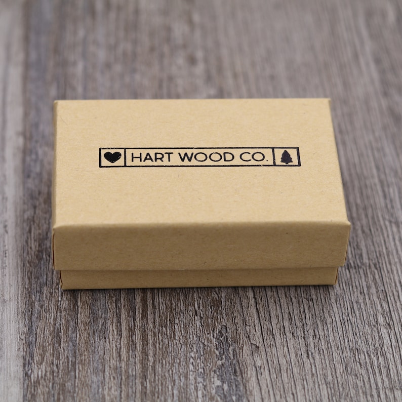 May include: A small, closed, rectangular cardboard box in a natural tan color. The box has a black stamp that reads "HART WOOD CO." with a heart and a tree symbol. The box is on a wooden surface.