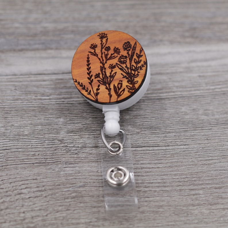 Puede incluir: Un portagafete retr&aacute;ctil de madera con un dise&ntilde;o floral. El portagafete es marr&oacute;n con un cord&oacute;n retr&aacute;ctil blanco y un clip plateado.