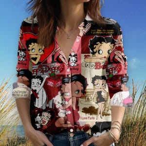 Peut inclure: Chemise boutonnée à manches longues avec un motif de collage d'illustrations de Betty Boop en rouge, rose et blanc. La chemise comprend du texte tel que "Betty Boop", "I Love Paris" et "Just like the moon, I go through phases". La chemise a un col en V.