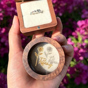 May include: A wooden ring box with a hinged lid and a circular base featuring a gold rose graphic and the name Tiffany. The open box reveals a silver ring with heart-shaped accents resting on a white velvet insert within the square compartment.