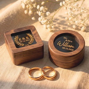 May include: Two dark wood ring boxes, one square and one round, featuring glass lids with gold text. The square box displays E & K 08.18.2025 with a laurel wreath, and the round box shows Mr. & Mrs. William 08.25.2026. Two gold bands sit in front.