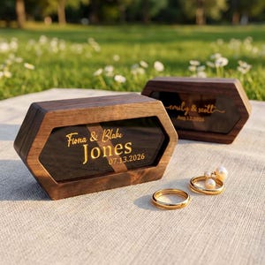 May include: Two hexagonal dark wood ring boxes with black acrylic lids. The front box features gold text reading Fiona & Blake Jones 07.13.2026. The back box displays gold text reading emily & scott Aug.12.2026. Two gold rings and pearl earrings.