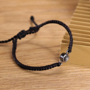 May include: A black string bracelet with a silver charm. The charm is round and has a dark gray center.