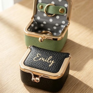 Könnte beinhalten: Zwei kleine, quadratische Schmuckkästchen mit goldfarbenen Rahmen und Verschlüssen. Eines ist schwarz mit dem Namen "Emily" in goldener Schrift. Das andere ist grün, offen und mit weißen Punkten gefüttert. Ein Ring ist in der grünen Box sichtbar.