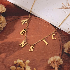 May include: A gold-colored necklace with the name "KENSIC" spelled out in individual letter pendants. The letters are suspended from a delicate chain. The necklace is displayed on a wooden surface with dried flowers.