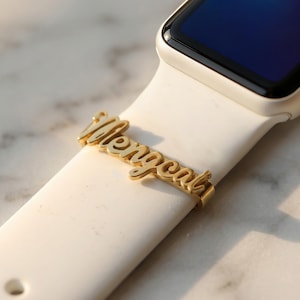 May include: Gold-colored watch band charm with the name "Mengcal" in a cursive font. The charm is attached to a white watch band. The watch face is black.