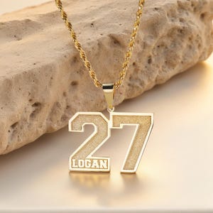 May include: Gold-colored pendant necklace with the number "27" and the name "LOGAN" in a rectangular box. The pendant hangs from a gold rope chain. The background is a light brown, textured surface.