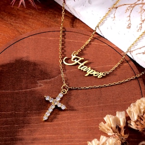 May include: A gold-colored layered necklace. The top layer features the name "Harper" in a cursive font. The bottom layer has a cross pendant adorned with clear gemstones. The necklace is displayed on a wooden surface.