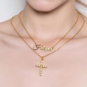 May include: A layered gold necklace featuring a cursive nameplate that reads "Harper" and a pendant with a cross design. The cross is adorned with sparkling, clear gemstones. The jewelry is displayed against a neutral background.