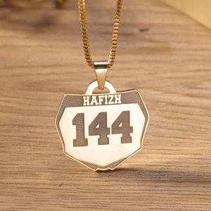 May include: Gold-coloured pendant necklace with a rectangular shield design. The pendant features the name "HAFIZH" above the number "144" in bold, black lettering. The necklace is displayed on a wooden surface.