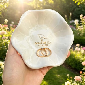 May include: A white ceramic ring dish with a scalloped edge, holding two gold rings. The dish is inscribed with "Cecilia UCLA 2026" and a graduation cap design. The dish is held in a hand, with a blurred garden background.