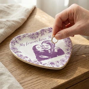 Peut inclure: Vide-poche en céramique en forme de cœur avec une bordure florale et un bord doré. Le plat présente une illustration violette de deux personnes s'embrassant et le texte "My forever hero, Mom". Une main tient une bague en or.