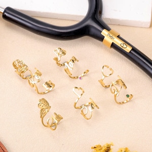 May include: Gold-colored stethoscope charms with various designs, including a flower, a heart, and a stethoscope. One charm has the name "Dukes" engraved. The charms are displayed on a beige surface.