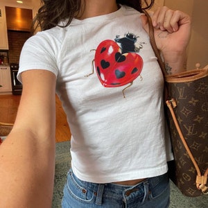 Pode incluir: Uma camiseta branca cropped com um gráfico de uma joaninha em um coração vermelho. A joaninha tem manchas pretas e uma cabeça preta. O coração tem corações pretos sobre ele.