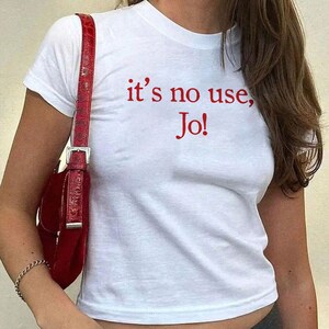 May include: A white short-sleeved t-shirt featuring the red text "it's no use, Jo!" printed on the chest. The shirt has a crew neckline and a cropped fit. A red textured shoulder bag with a silver buckle is worn over the shoulder.