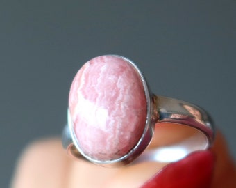 Puede incluir: Un anillo de plata con una piedra ovalada de color rosa y blanco. El anillo está en un dedo con esmalte de uñas rojo.