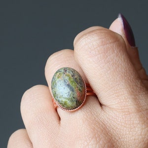 Puede incluir: Un anillo de cobre con una piedra ovalada verde y marrón. El anillo está en un dedo.