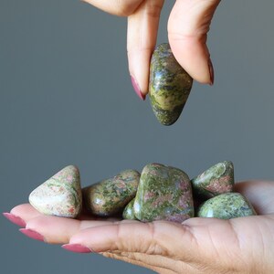 May include: A hand holding a collection of green and pink moss agate tumbled stones. The stones are smooth and polished, with a variety of patterns and colors.