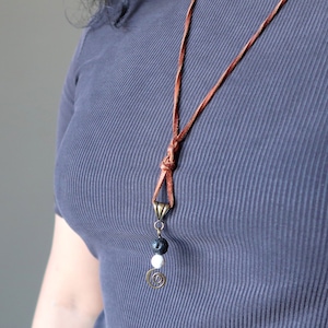 Puede incluir: Un collar de cordón de cuero marrón con un colgante de latón y tres cuentas. Las cuentas son negras, blancas y negras. El colgante tiene un diseño en espiral.