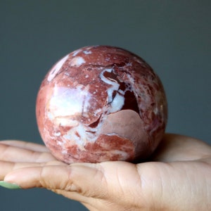 Può includere: Una sfera di marmo rosso e bianco, lucidata a una finitura liscia. La sfera è tenuta in una mano, mostrando i suoi intricati motivi e la sua bellezza naturale.