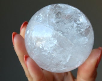 May include: A clear quartz crystal sphere, held in a hand with red nail polish. The sphere is translucent with internal fractures, reflecting light. The background is a muted gray.