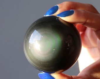 May include: A black obsidian sphere with iridescent rainbow colors, reflecting light. The sphere is being held in a hand with blue nail polish.