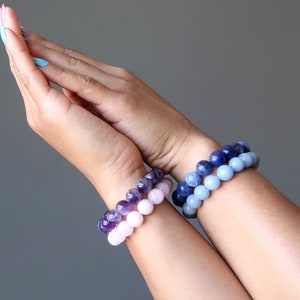 May include: Two bracelets made of gemstone beads. The top bracelet is made of light pink and purple beads. The bottom bracelet is made of light blue beads.