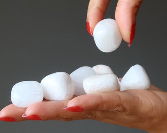May include: A hand holding a collection of white selenite tumbled stones. The stones are smooth and polished, with a slightly translucent appearance.