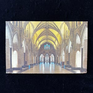 Op de afbeelding: Een vintage ansichtkaart met het interieur van een kathedraal. De afbeelding toont een lange, gewelfde gang met zuilen en een hoog, gewelfd plafond. Het kleurenpalet wordt gedomineerd door warme tinten beige, bruin en geel, met een vleugje blauw van het glas-in-loodraam.
