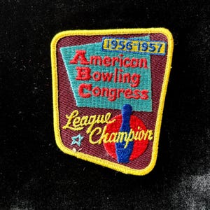 May include: A vintage patch with a maroon background and a yellow border. The patch features the text "American Bowling Congress" in red, "League Champion" in yellow, and the years "1956-1957". A blue bowling pin is also visible.