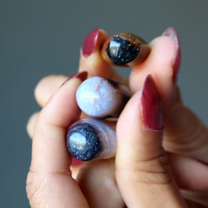 May include: Three agate gemstone rollers held in a hand. The rollers are a light blue, a dark brown with black swirls, and a black with white swirls.