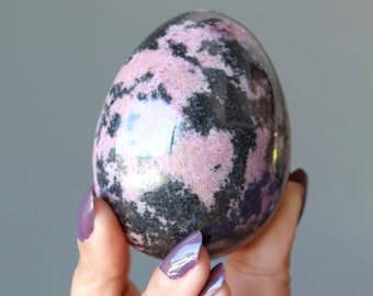 May include: A polished pink and black rhodonite egg. The egg is smooth and has a mottled appearance.