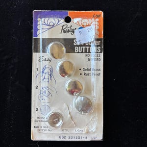 May include: A vintage package of Prestige S.S. Snapper Buttons. The package contains five silver-colored, round buttons. The card reads "Easy" with instructions and "Solid Brass" and "Rust Proof". The package is marked "Made in U.S.A." and the buttons are 3/4 inch in diameter.
