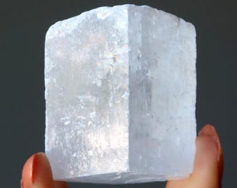 Może przedstawiać: Półprzezroczysty, biały, prostokątny blok kryształu selenitu o włóknistej, warstwowej strukturze. Okaz minerału wykazuje naturalną strukturę krystaliczną z subtelnymi opalizującymi refleksami na gładkich, płaskich powierzchniach.