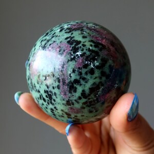 Może przedstawiać: Zielona, czarna i różowa kula z kamienia szlachetnego. Kula jest gładka i wypolerowana, z nakrapianym wzorem.