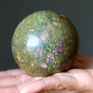 Peut inclure: Une pierre sphérique polie présentant une base de couleur vert marbré avec des inclusions cristallines incrustées dans des tons magenta et rose foncé. La surface de la sphère est lisse et réfléchissante, révélant un motif minéral naturel.
