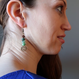 May include: A pair of green and brown gemstone earrings with a spiral design. The earrings are hanging from a person's ear.
