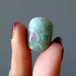 Peut inclure: Une pierre roulée verte et rose avec une surface lisse et arrondie. La pierre présente un motif unique d'inclusions roses et blanches.