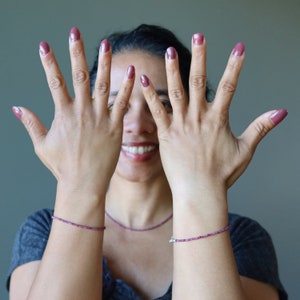 Puede incluir: Manos con uñas pintadas en un tono rosa intenso, con pulsera y collar de cuentas a juego. El fondo es gris liso.